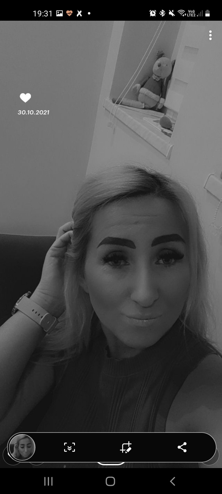 Portret kobiety z blond włosami i zegarkiem na ręce, w tle półka z pluszowym misiem, zdjęcie czarno-białe.