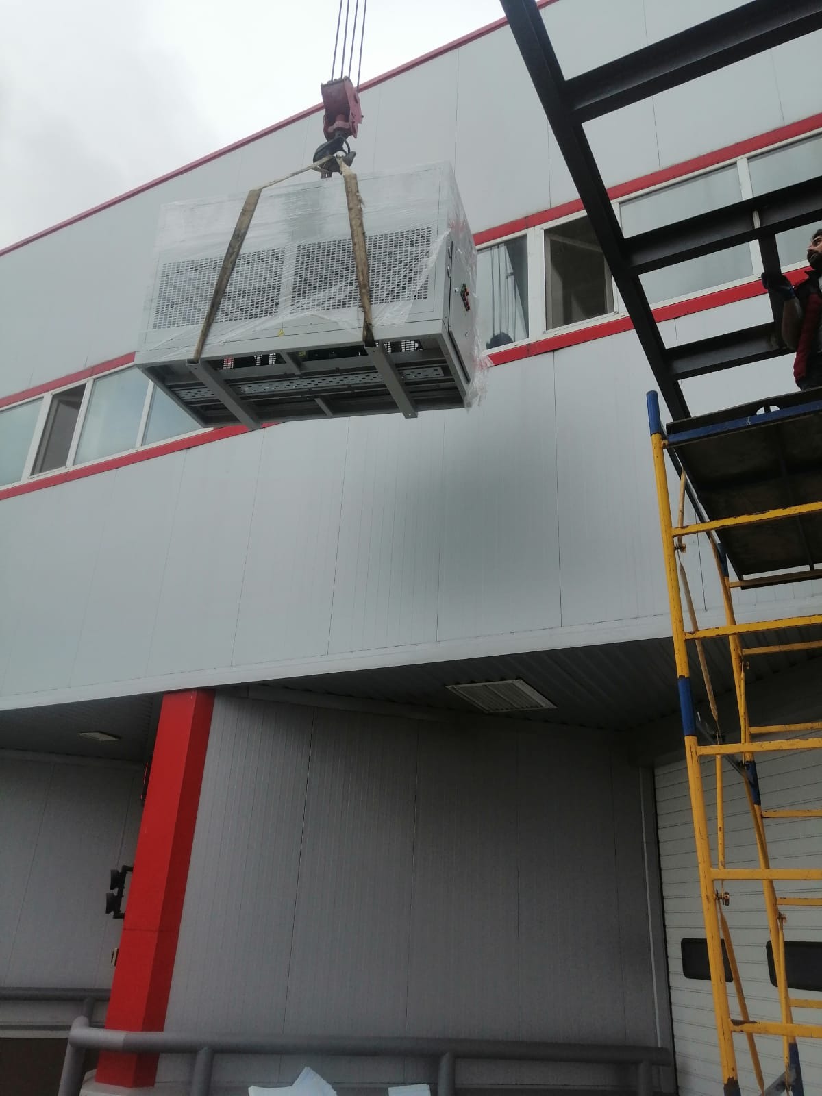 Podnoszenie klimatyzatora przemysłowego w folii ochronnej za pomocą dźwigu na tle hali z czerwonymi akcentami i żółtą drabiną.