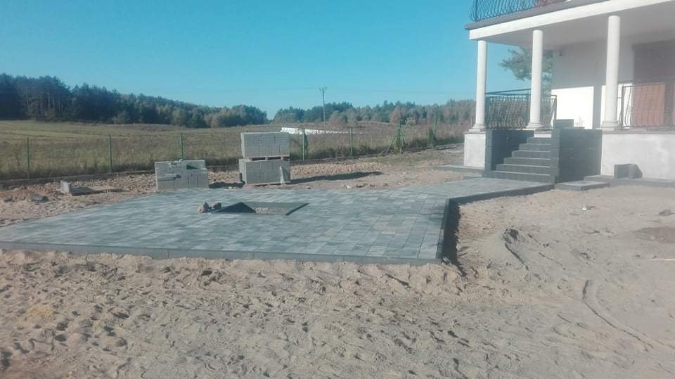 Fragment podwórza w trakcie prac: ułożona szara kostka brukowa tworząca taras i ścieżkę prowadzącą do schodów wejściowych domu z kolumnami, obok sterta kostki do ułożenia, w tle zielone pole i las...