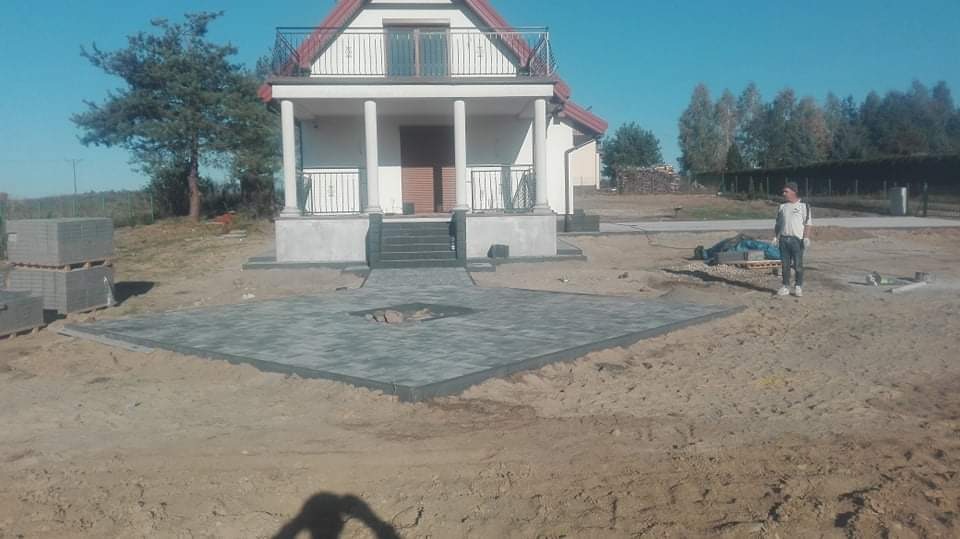 Częściowo ułożona, kwadratowa powierzchnia z szarej kostki brukowej z otworem pośrodku, na tle domu z kolumnami i pracownika w oddali.