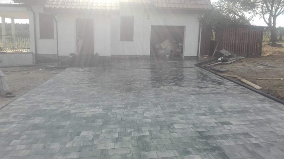 Świeżo ułożona, szara kostka brukowa na podjeździe przed domem z garażem, widoczne pozostałości piasku i narzędzia na obrzeżach.