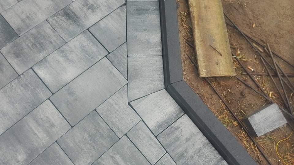 Ułożona szara kostka brukowa z ciemnoszarą obwódką, widoczne wystające pręty zbrojeniowe i fragment deski na ziemi.