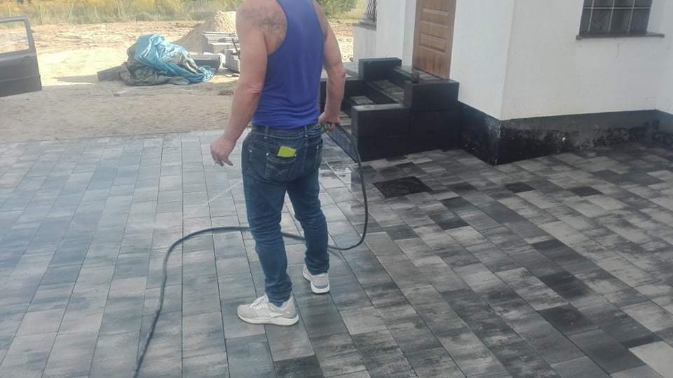Mężczyzna w jeansach i niebieskim podkoszulku myje wodą pod ciśnieniem nowo ułożoną, szaro-czarną kostkę brukową przed domem. W tle widoczne schody wejściowe z ciemnego kamienia i przygotowany...