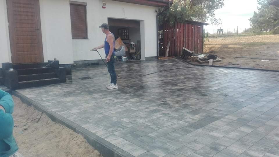 Pracownik spryskuje wodą nowo ułożoną, szarą kostkę brukową na podjeździe przed domem z drewnianymi schodami wejściowymi i garażem z boku.