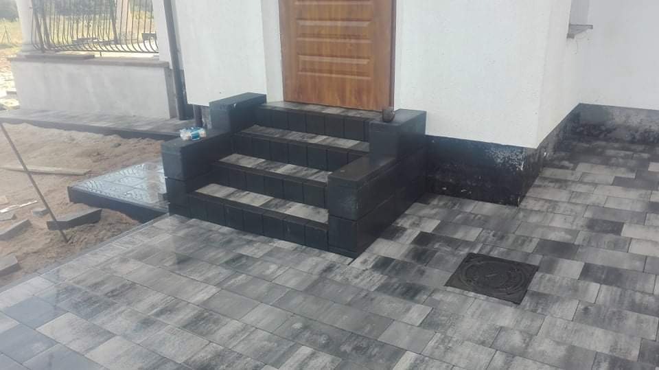 Nowe, trzystopniowe schody wejściowe z ciemnej kostki brukowej prowadzące do drewnianych drzwi, otoczone świeżo ułożonym chodnikiem z szarej kostki brukowej.