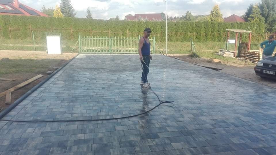 Pracownik myje świeżo ułożoną kostkę brukową na podjeździe, widoczny wąż ciśnieniowy i mokra powierzchnia kostki, w tle ogrodzenie posesji i samochód.