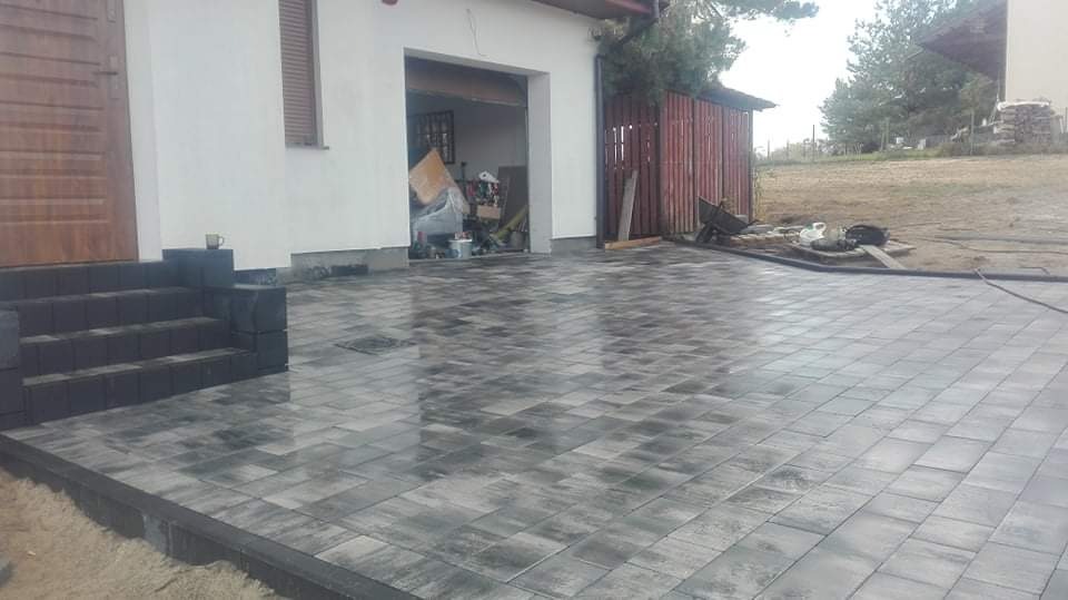 Ułożona szara kostka brukowa przed domem z garażem, widoczne schody wejściowe z kostki i narzędzia budowlane w tle.