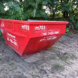 Czerwony kontener na gruz z białymi napisami 'm703' i 'NIE ZASTAWIAC' oraz numerem telefonu, stojący na ziemi z widocznymi śladami opon, w tle drzewa.