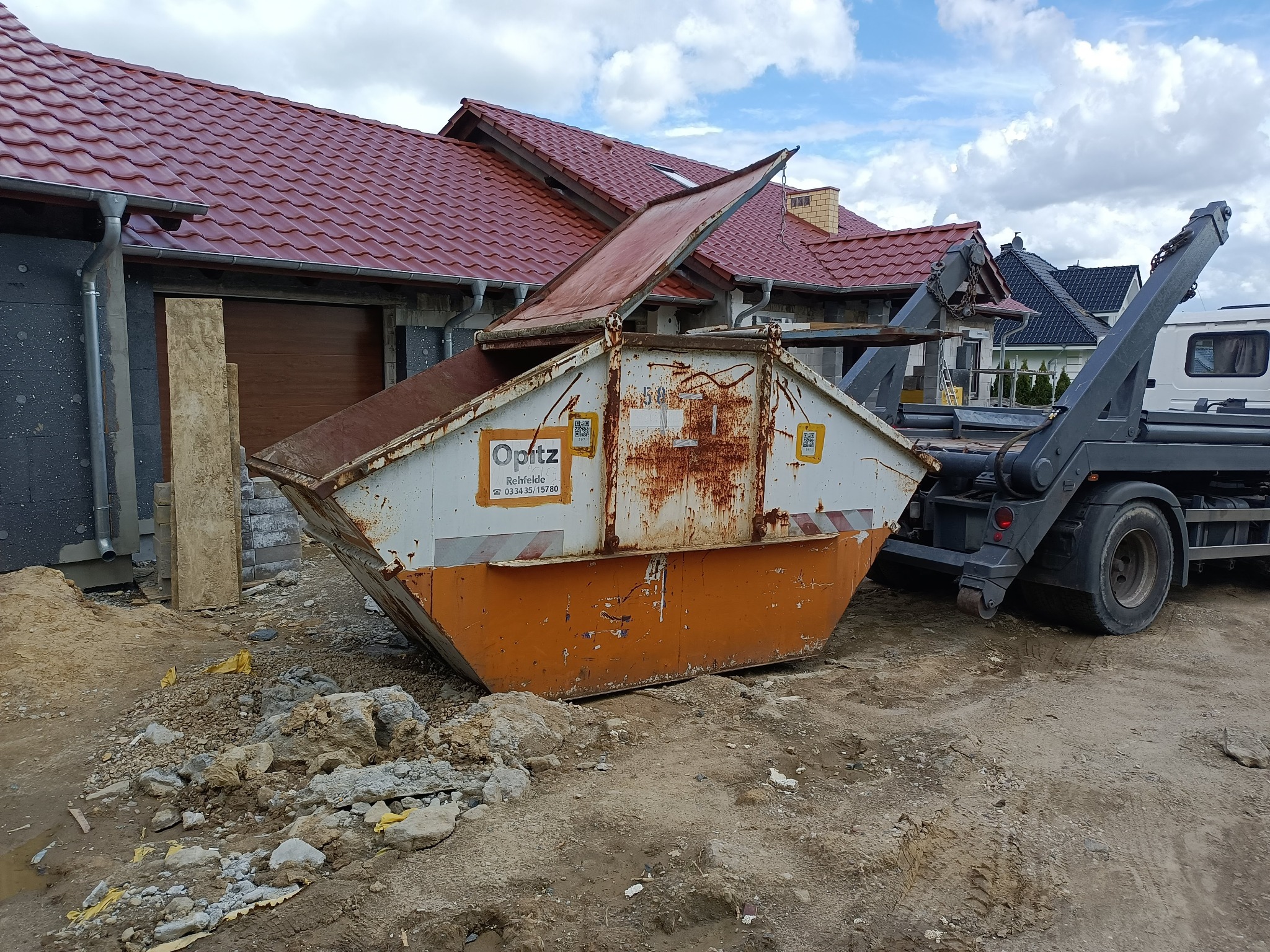 Pomarańczowo-biały, częściowo zardzewiały kontener na gruz z otwartą klapą, stojący na ziemi z gruzem, obok szarej ciężarówki z ramieniem podnoszącym. W tle niedokończony dom z czerwoną dachówką...