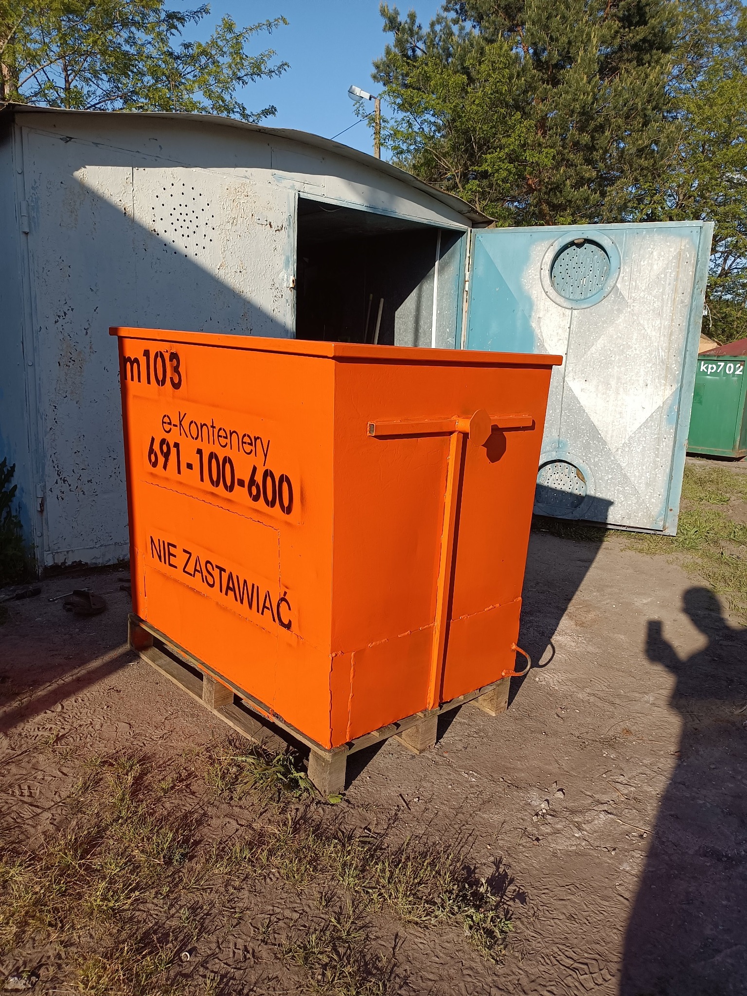 Pomarańczowy kontener na gruz z czarnym napisem 'm103 e-kontenery 691-100-600 NIE ZASTAWIAĆ' stoi na drewnianej palecie przed otwartym, blaszanym garażem.