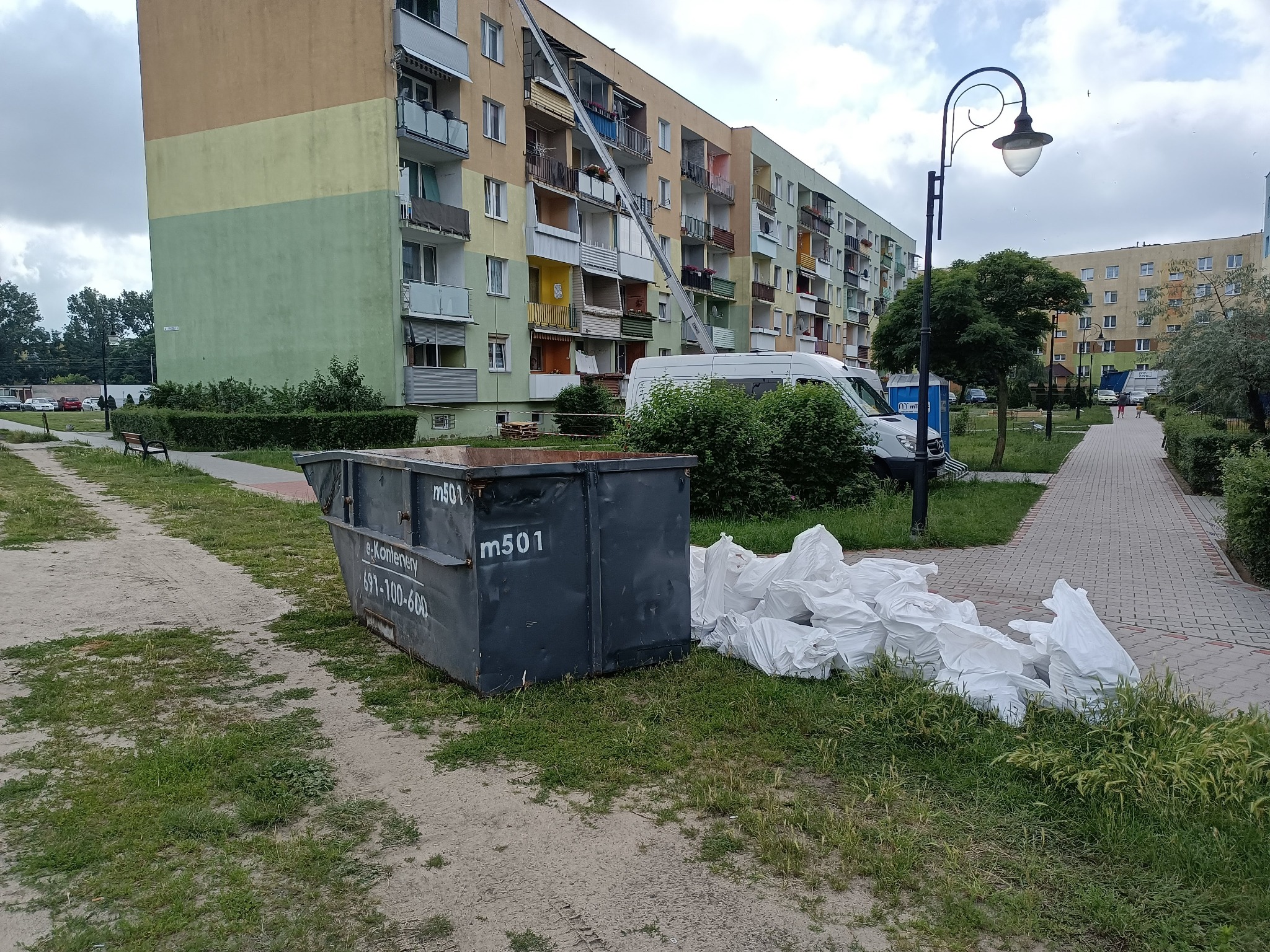 Metalowy kontener m501 obok sterty białych worków na gruz na trawie, z blokiem mieszkalnym i podnośnikiem meblowym w tle.