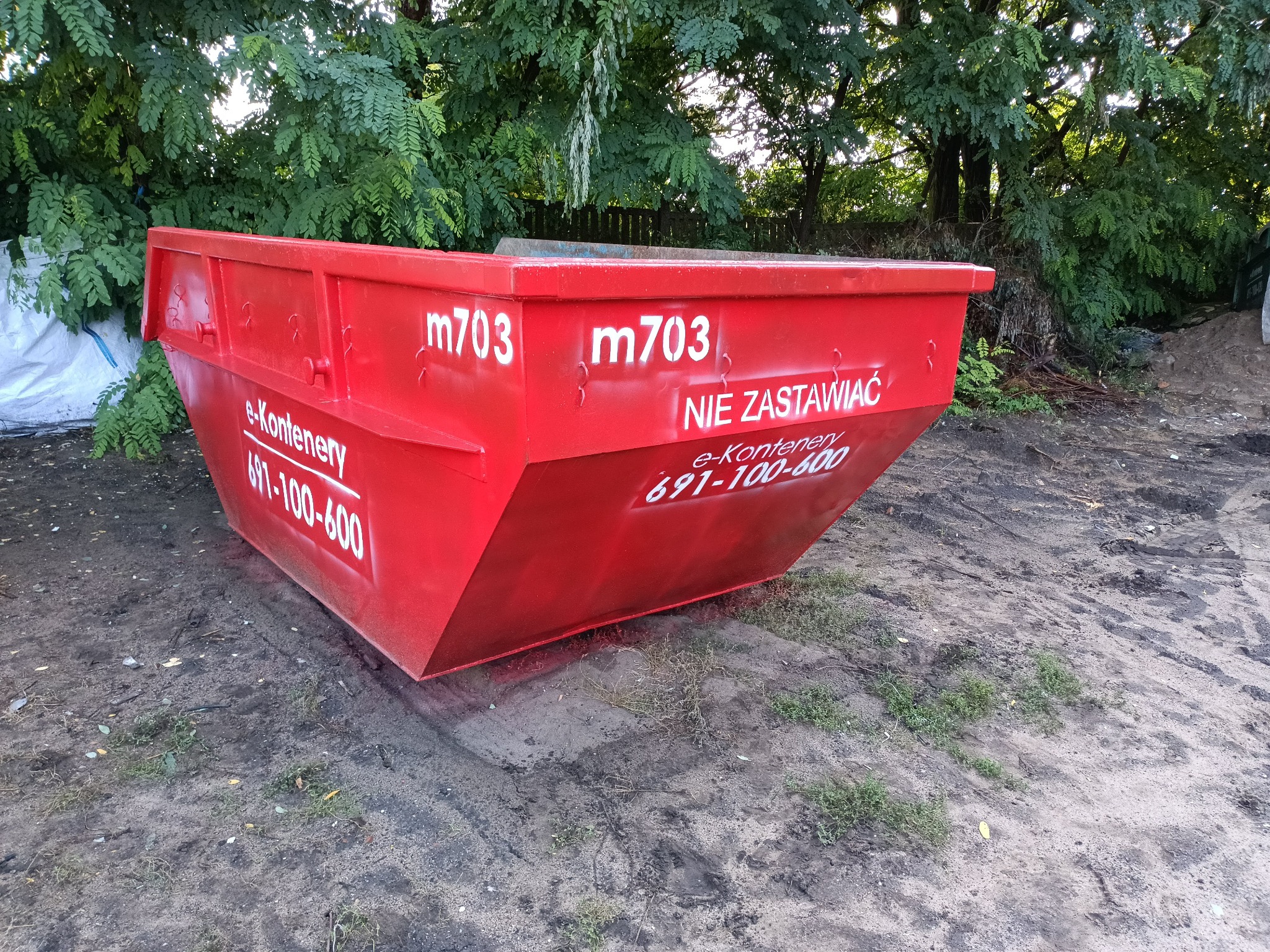 Czerwony kontener na gruz z białymi napisami 'm703' i 'NIE ZASTAWIAC' oraz numerem telefonu, stojący na ziemi z widocznymi śladami opon, w tle drzewa.