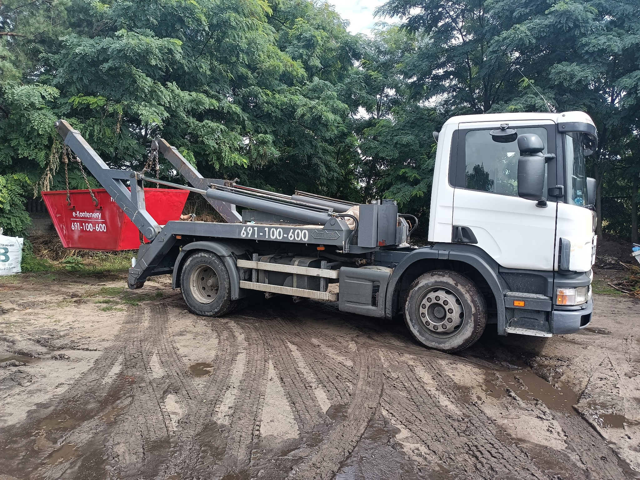 Ciężarówka z hakiem i czerwonym kontenerem na gruz, stojąca na błotnistym terenie z widocznymi śladami opon, drzewa w tle.
