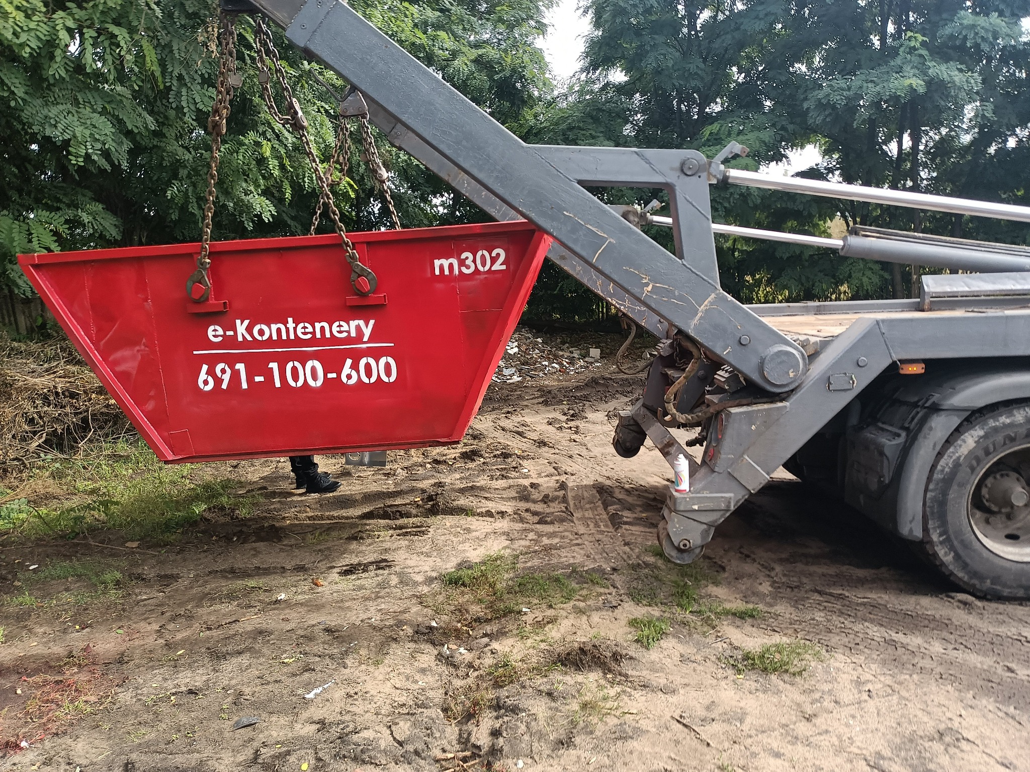 Czerwony kontener na gruz z napisem 'e-Kontenery' i numerem telefonu, podnoszony przez hakowy system załadunku na tle drzew i ziemi.