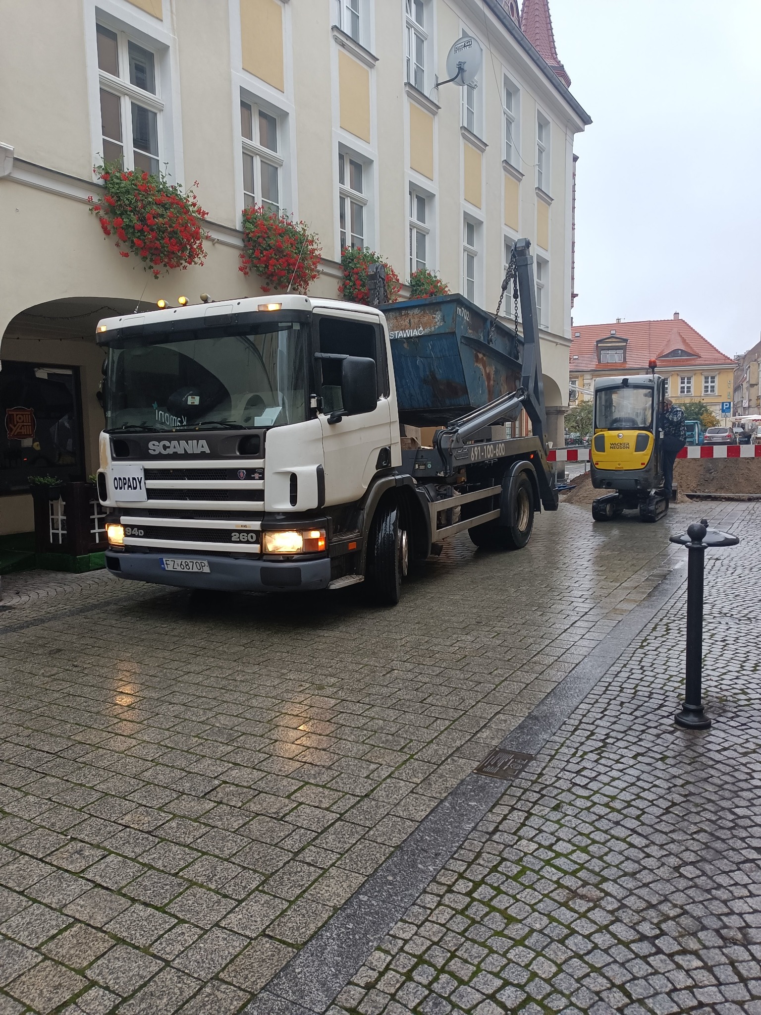Ciężarówka Scania z kontenerem na odpady na brukowanej ulicy starego miasta; widoczny żółty mini-koparka Wacker Neuson w tle, obok wykopu z piaskiem.