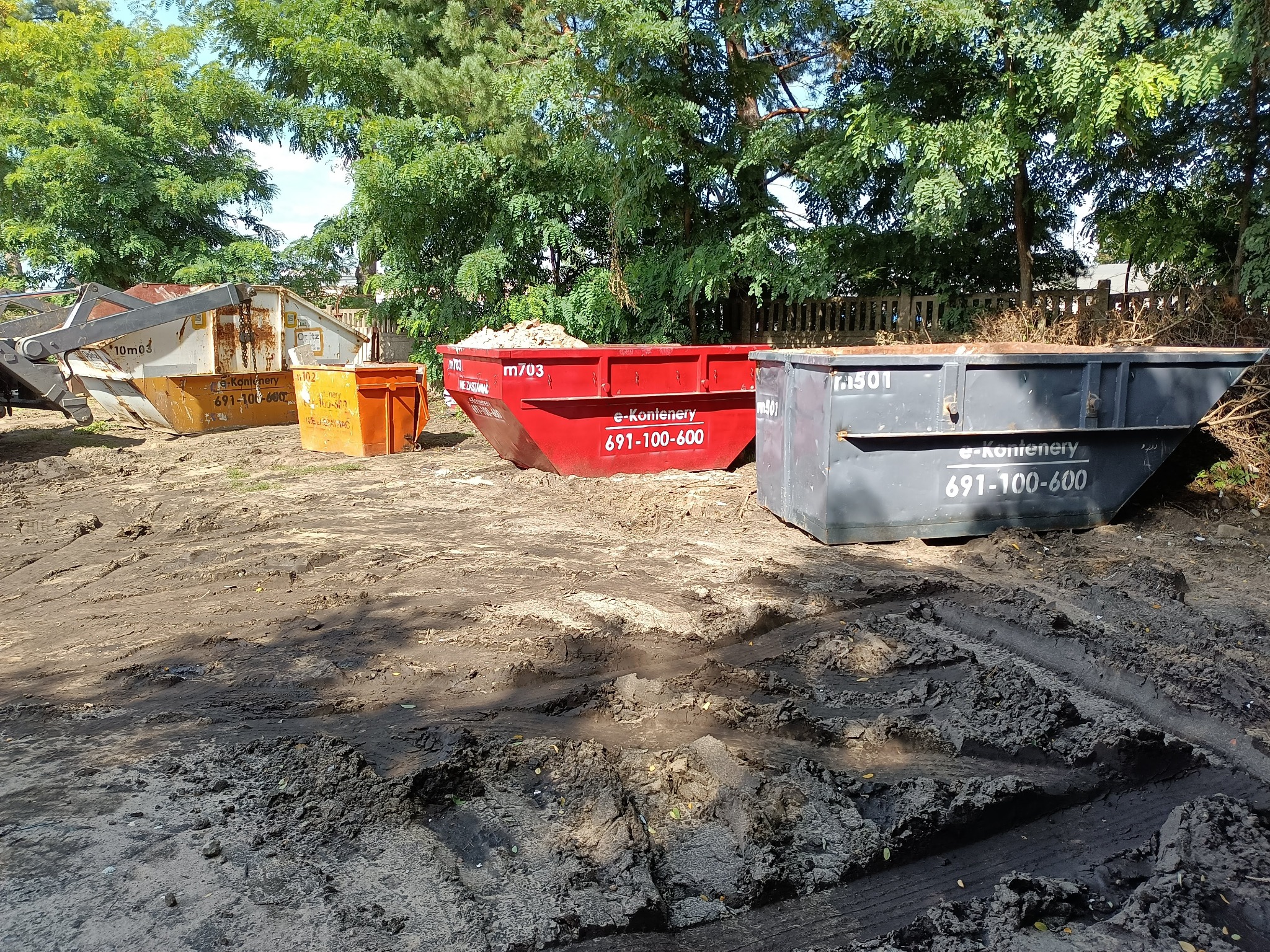 Cztery kontenery na gruz różnych kolorów (biały, pomarańczowy, czerwony, szary) stojące na ubłoconym terenie pod drzewami, widoczne ślady opon na ziemi.