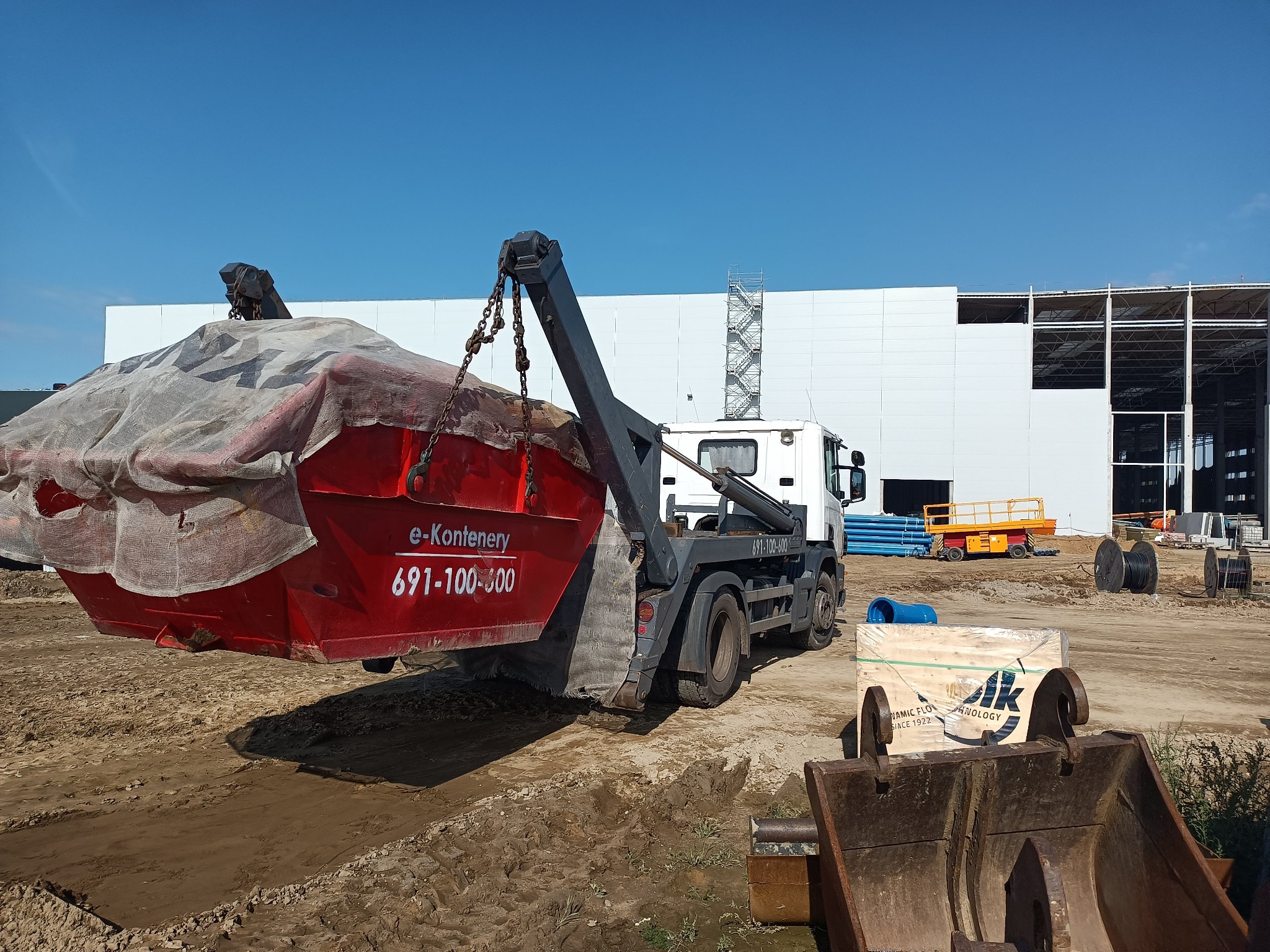 Ciężarówka z hakiem podnosi czerwony, zakryty kontener na gruz na placu budowy, w tle biały budynek w trakcie budowy.