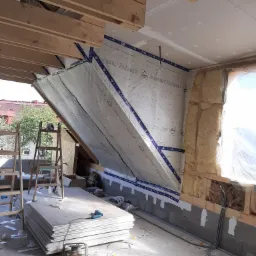 Poddasze w trakcie izolacji: widoczna wełna mineralna, folia paroizolacyjna Ampack Vario 3, płyty karton-gips, drewniana konstrukcja szkieletowa, drabina oparta o ścianę i stos płyt G-K na podłodze.