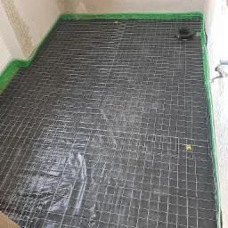 Przygotowanie podłoża pod ogrzewanie podłogowe w łazience: czarna folia izolacyjna, siatka zbrojeniowa i taśma uszczelniająca przy ścianach.