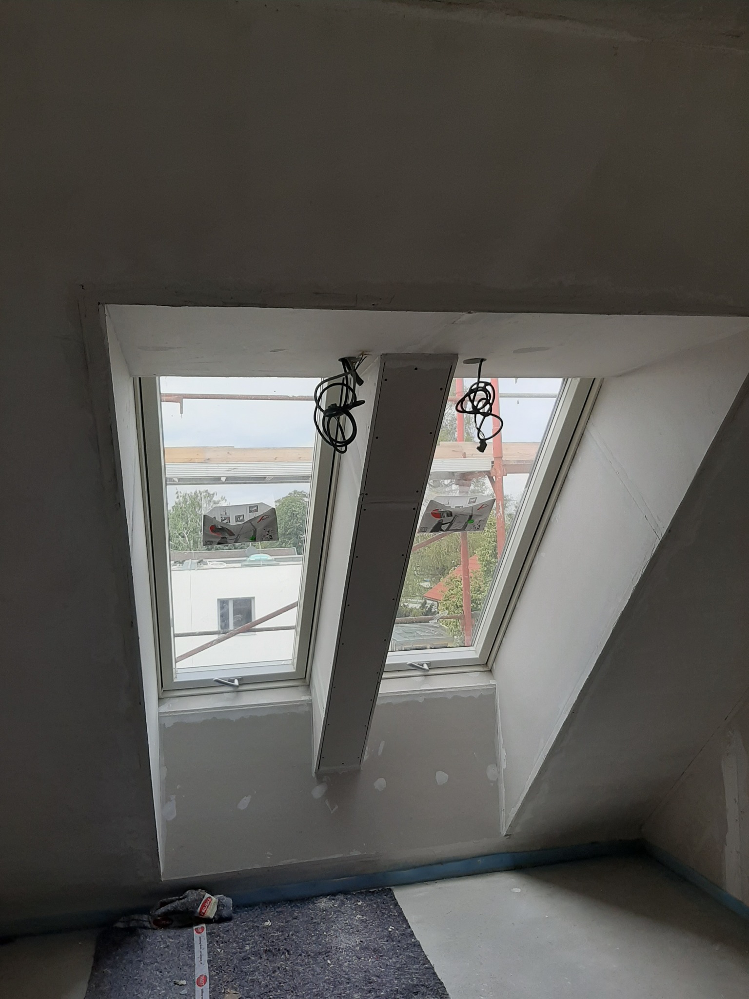 Poddasze w trakcie wykańczania z dwoma oknami dachowymi, widoczne kable zwisające z sufitu i fragment rusztowania za oknem, ściany przygotowane do malowania, widoczne ślady szpachlowania.