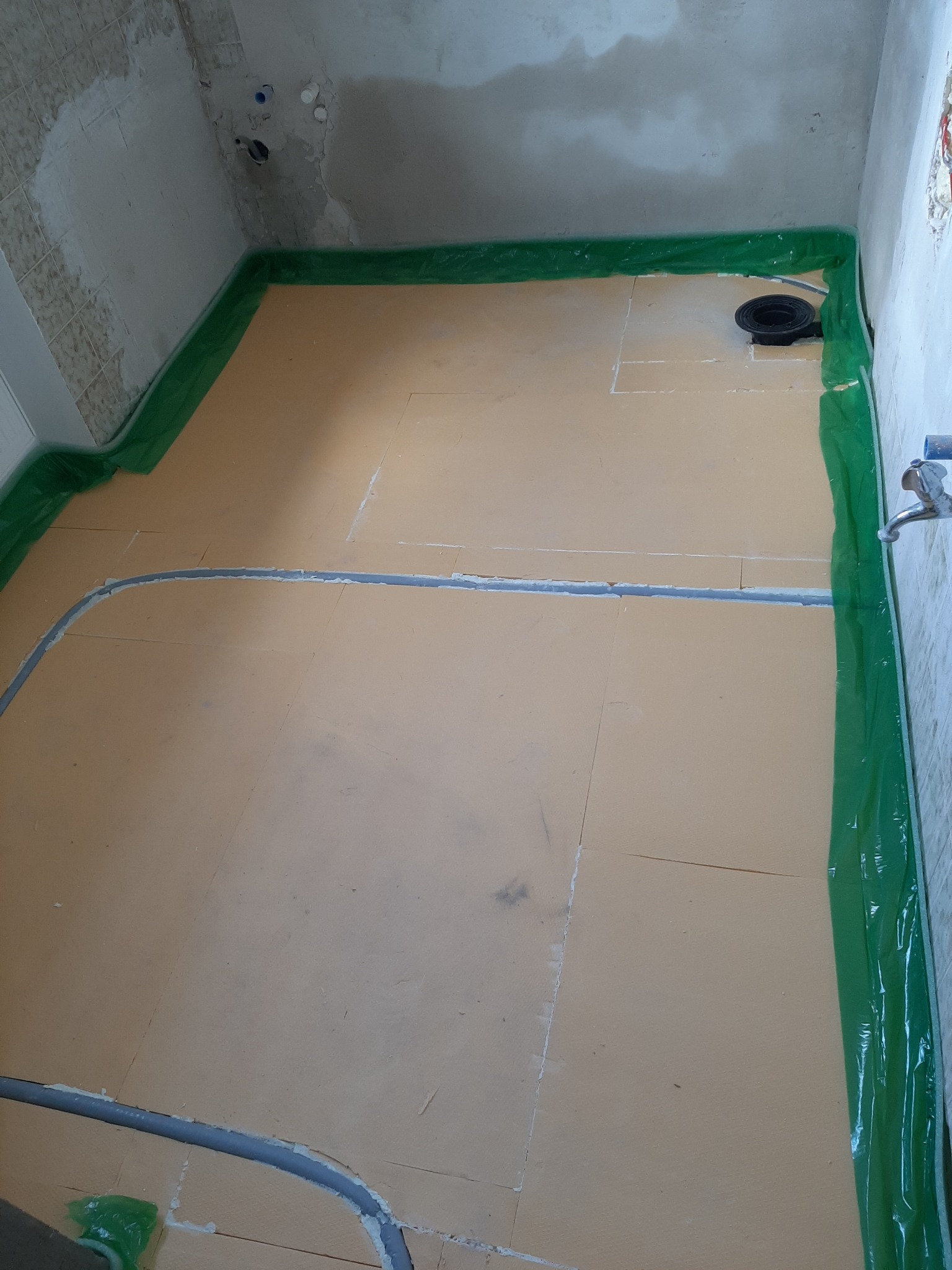 Przygotowanie podłoża w łazience: żółte płyty izolacyjne ułożone na podłodze, zabezpieczone folią wzdłuż ścian, widoczny odpływ liniowy i rury instalacji wodnej.