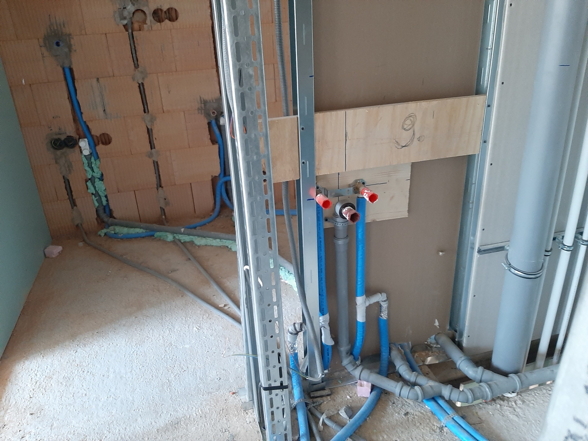 Wnętrze w trakcie prac hydraulicznych: niebieskie rury instalacji wodnej i szare rury kanalizacyjne prowadzone wzdłuż ściany z cegły i ściany z płyt kartonowo-gipsowych, metalowe profile...