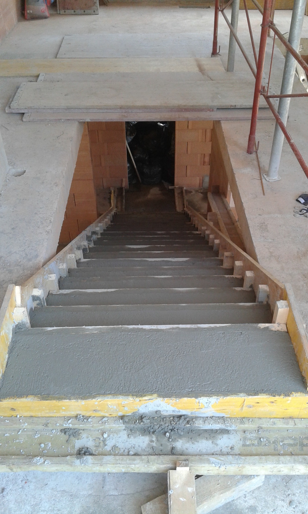 Surowe schody betonowe w szalunku drewnianym, w trakcie budowy, widok z góry na dół, z widocznymi cegłami i rusztowaniem w tle.