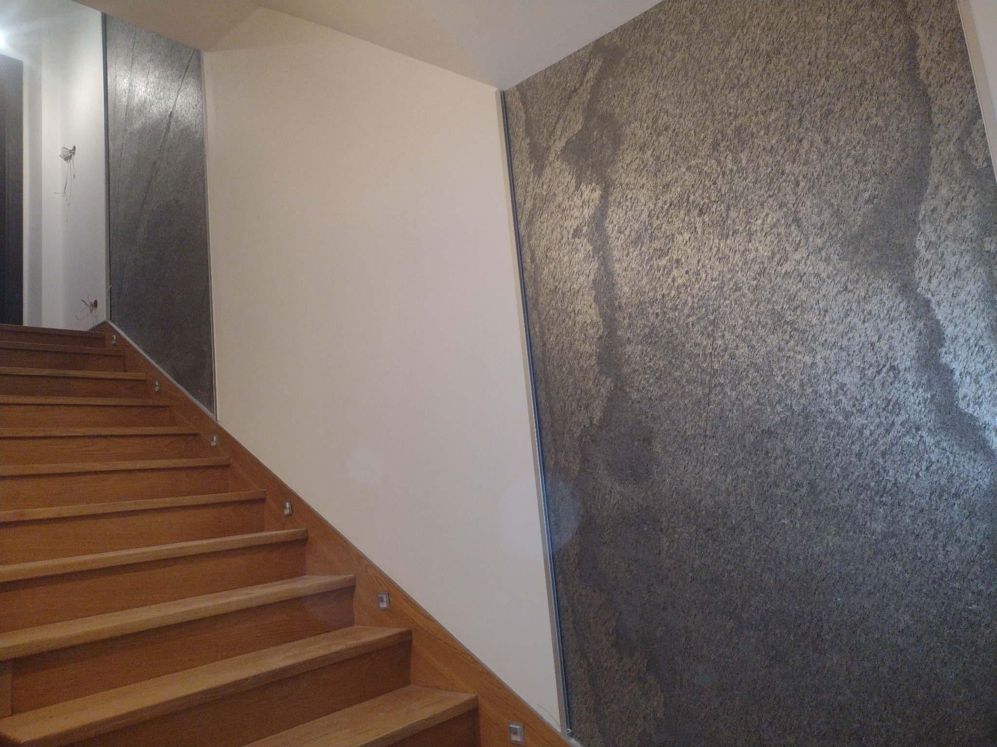 Klatka schodowa z drewnianymi schodami i ścianą pokrytą dekoracyjną szarą fakturą przypominającą kamień, sąsiadującą z gładką, białą ścianą.