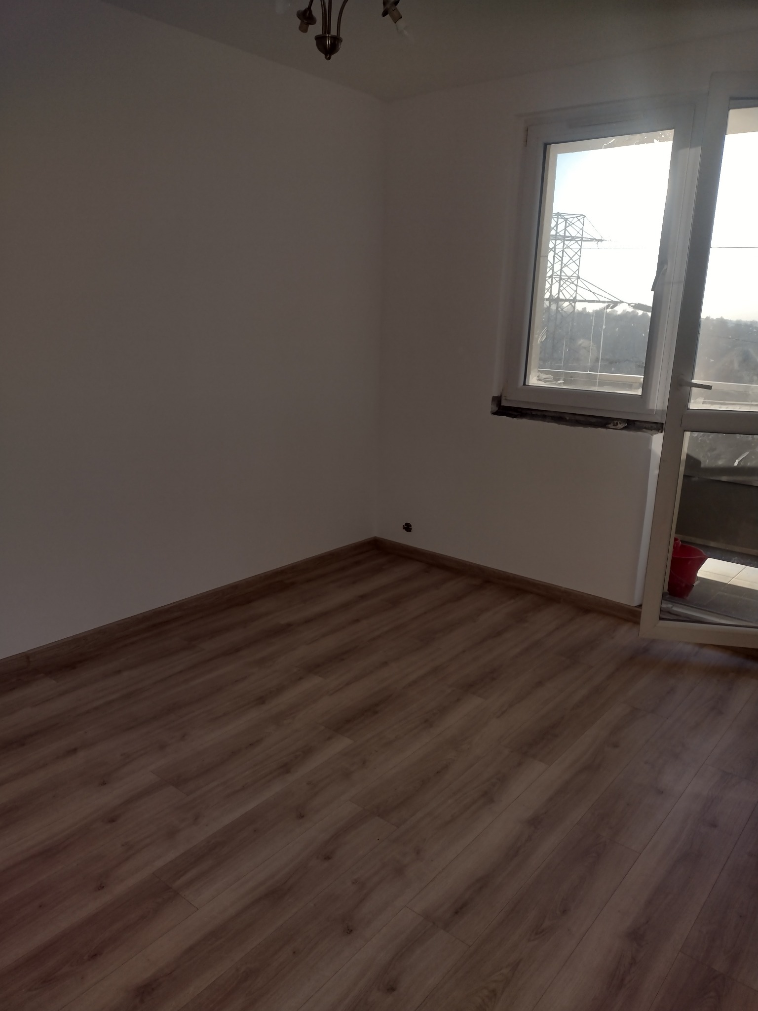 Pusty pokój z panelami podłogowymi w odcieniach brązu, świeżo pomalowanymi białymi ścianami i oknem balkonowym z widokiem na linię wysokiego napięcia.