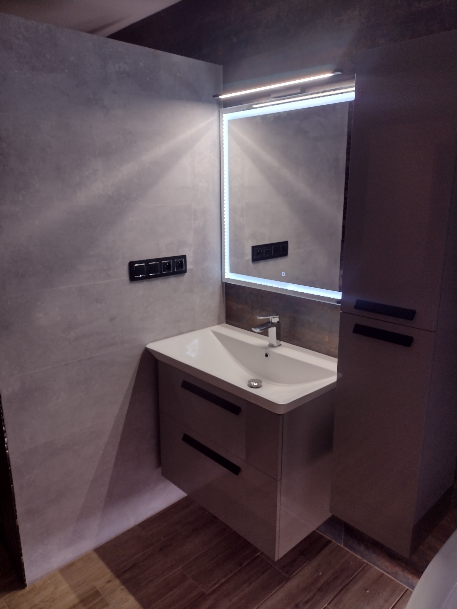 Nowoczesna łazienka z szarą płytką imitującą beton, podświetlanym lustrem nad białą szafką z umywalką i ciemnymi uchwytami, oraz brązową podłogą z paneli.