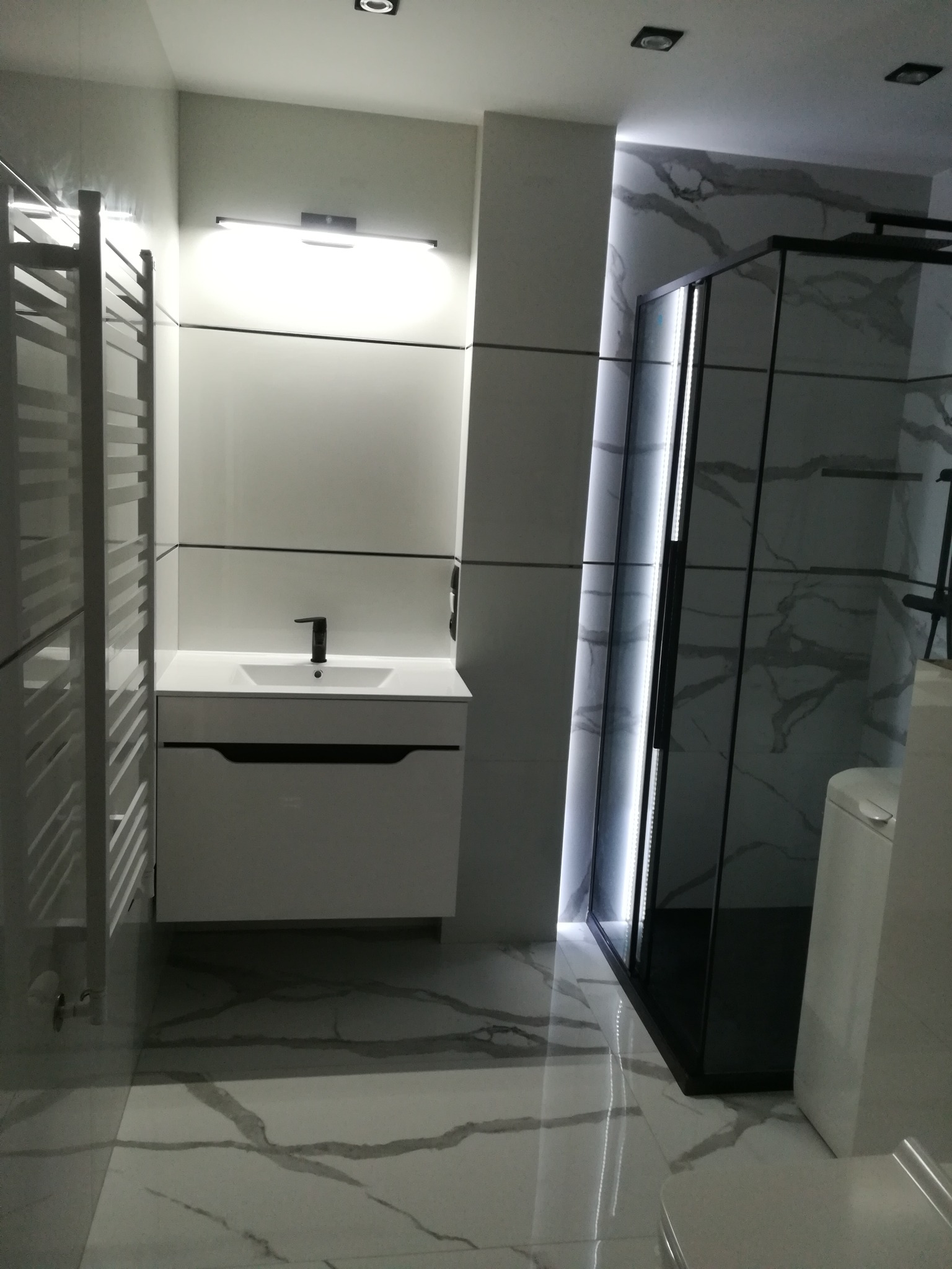 Nowoczesna łazienka z białymi płytkami imitującymi marmur, czarną kabiną prysznicową z podświetleniem LED, umywalką z szafką i grzejnikiem drabinkowym.