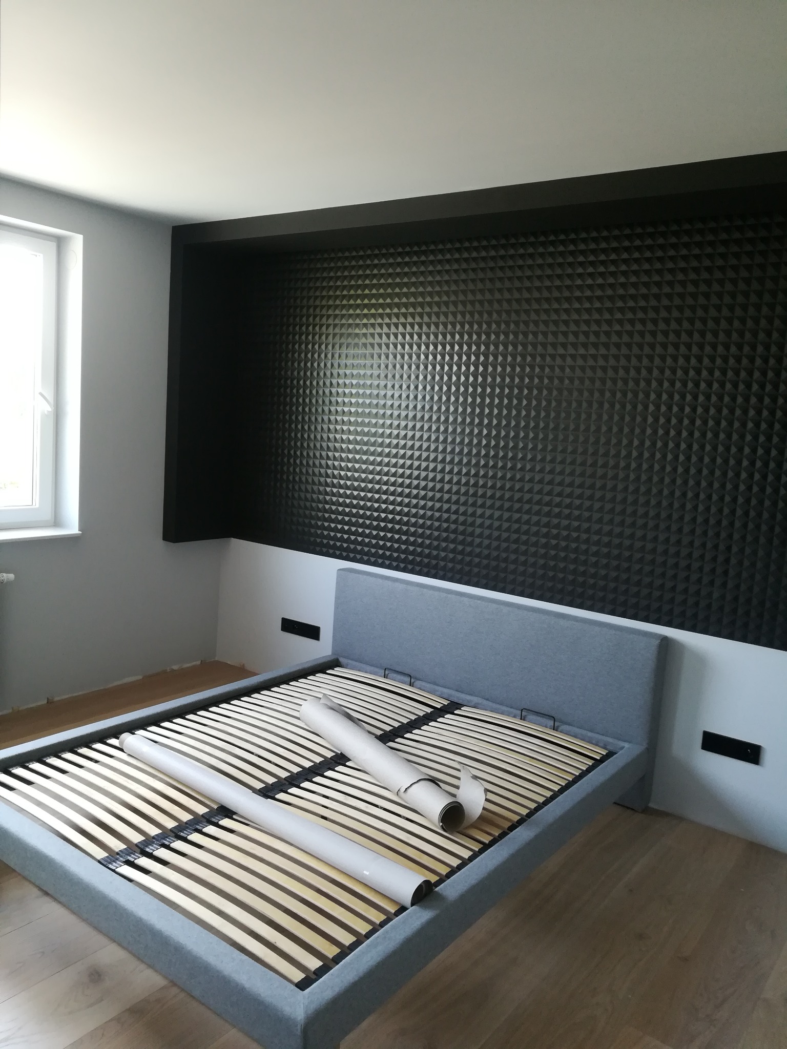 Szary stelaż łóżka z listwami i dwoma rulonami papieru, na tle ściany z ciemnymi panelami akustycznymi w pokoju z szarą ścianą i drewnianą podłogą.