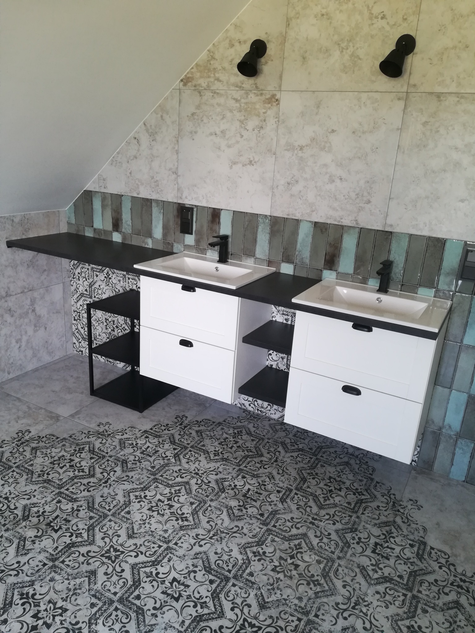 Dwie białe szafki pod umywalki z czarnymi blatami i armaturą w łazience na poddaszu, wzorzysta podłoga, dekoracyjne płytki na ścianach.