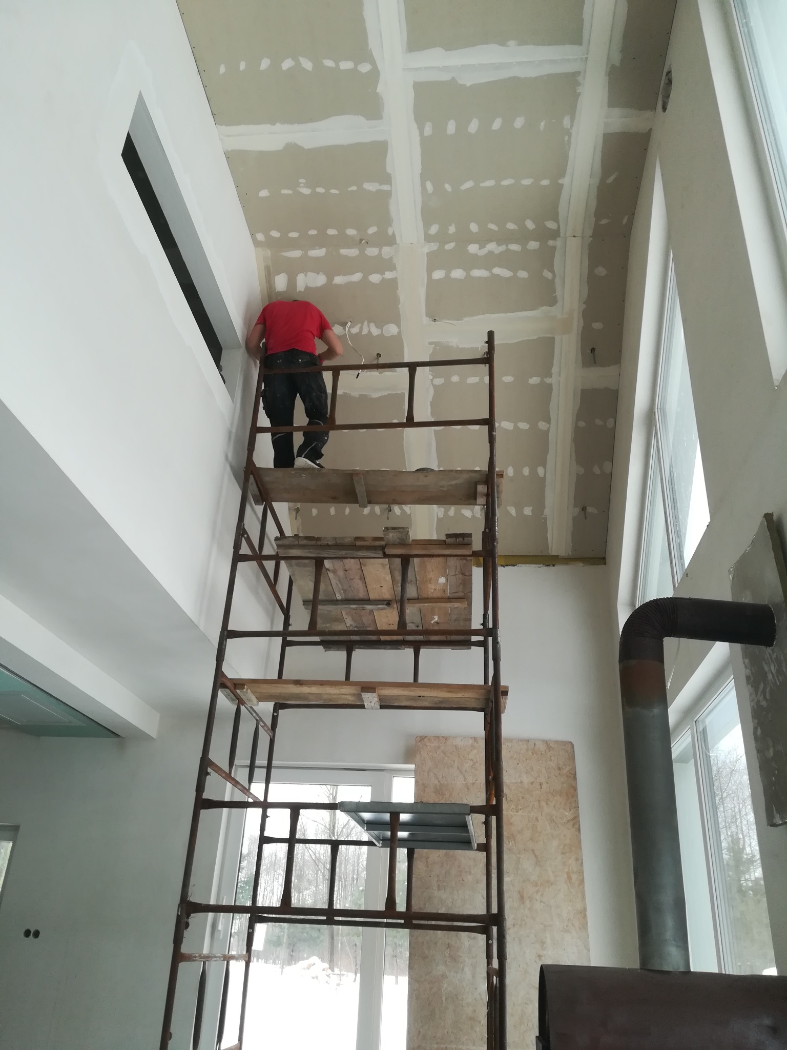 Pracownik na rusztowaniu montuje płyty na suficie w nowym, wysokim pomieszczeniu z dużymi oknami, widoczne łączenia płyt wypełnione masą.