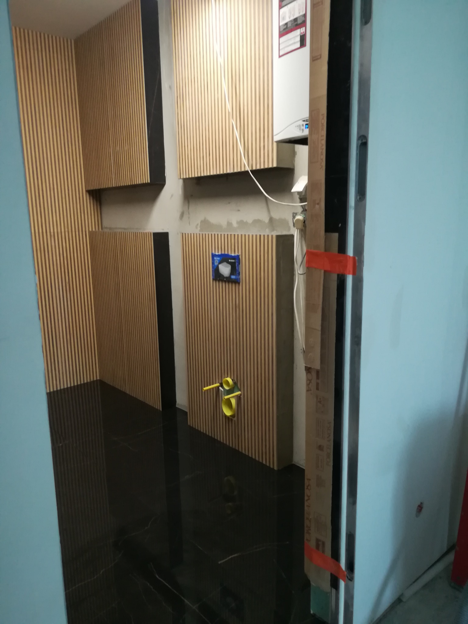 Wnętrze łazienki w trakcie wykańczania, widoczne panele dekoracyjne drewniane z czarnymi wstawkami na ścianach, czarna, błyszcząca podłoga, instalacja sanitarna w trakcie montażu, widoczny bojler.
