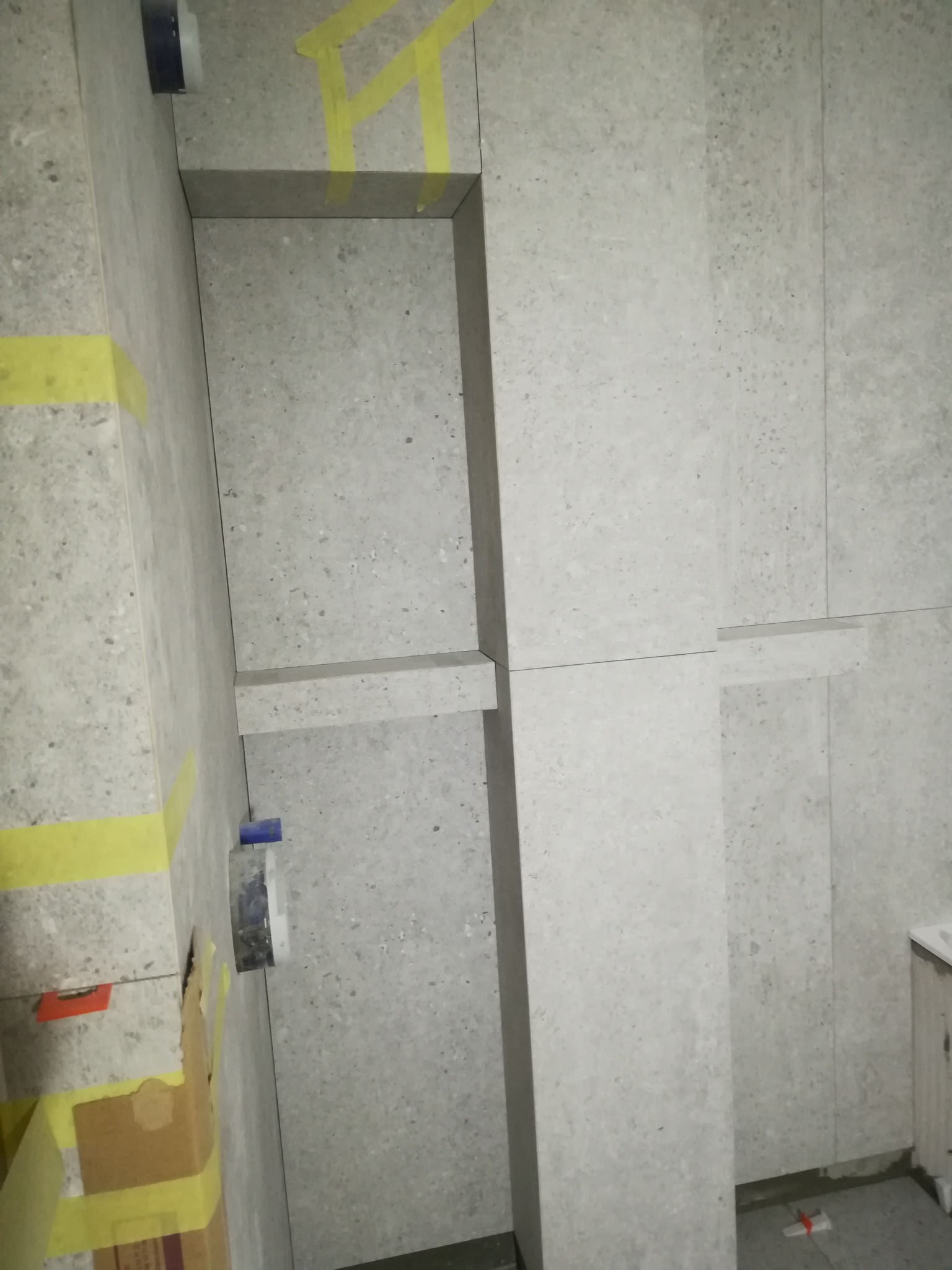 Wnętrze łazienki w trakcie prac wykończeniowych, widoczne płytki imitujące kamień, żółta taśma malarska zabezpieczająca krawędzie, oraz elementy instalacji sanitarnej.