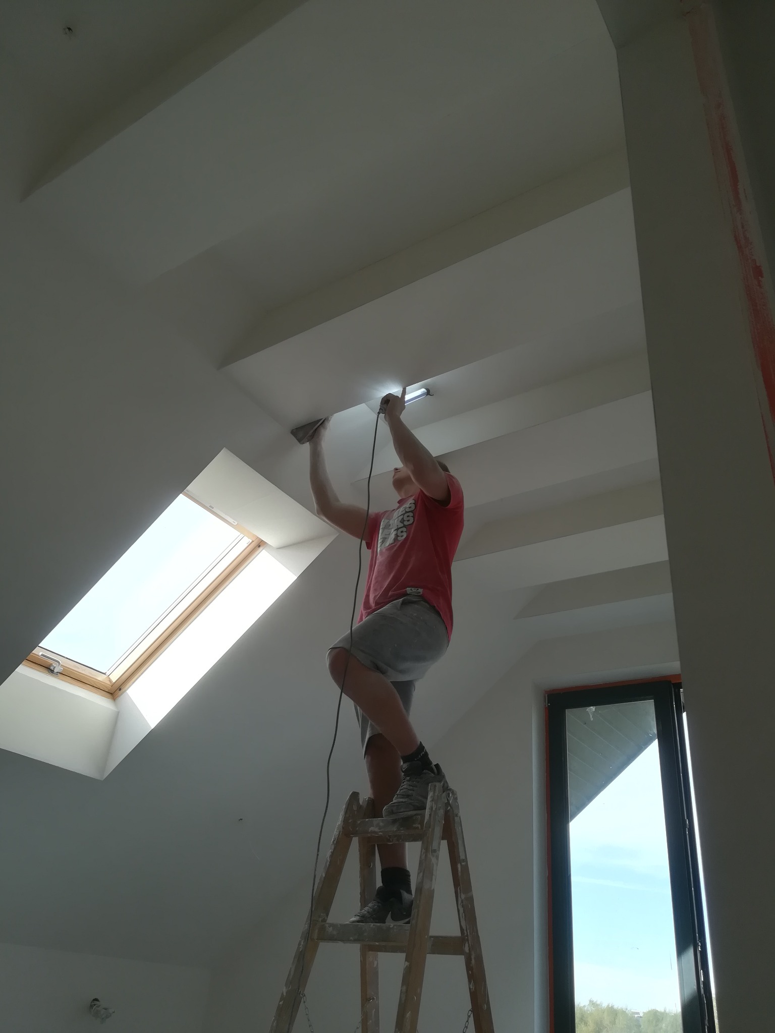 Pracownik na drabinie szlifuje połączenie płyty na skośnym suficie poddasza z oknem dachowym, widoczne ślady po szpachlowaniu.
