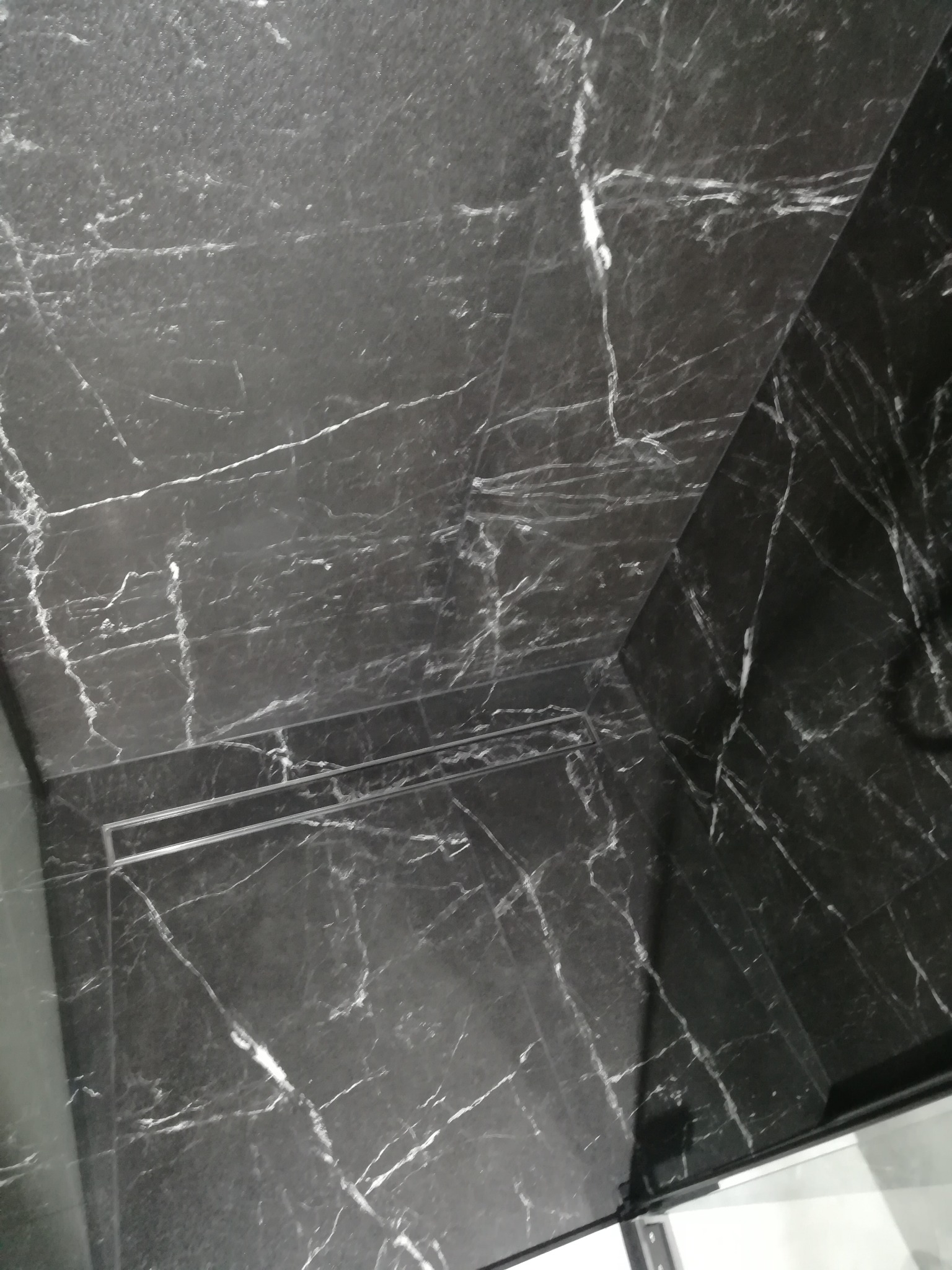 Wykończona kabina prysznicowa z czarnych płytek imitujących marmur z widocznym odpływem liniowym.