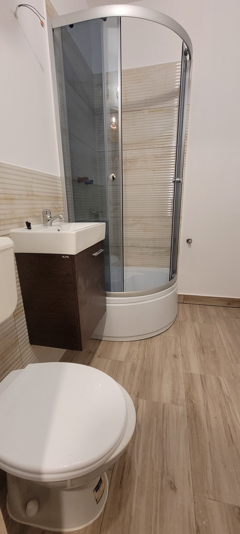 Ukończona łazienka z kabiną prysznicową narożną, umywalką z szafką w kolorze ciemnego drewna oraz toaletą, wykończona płytkami imitującymi drewno na podłodze i ścianach.