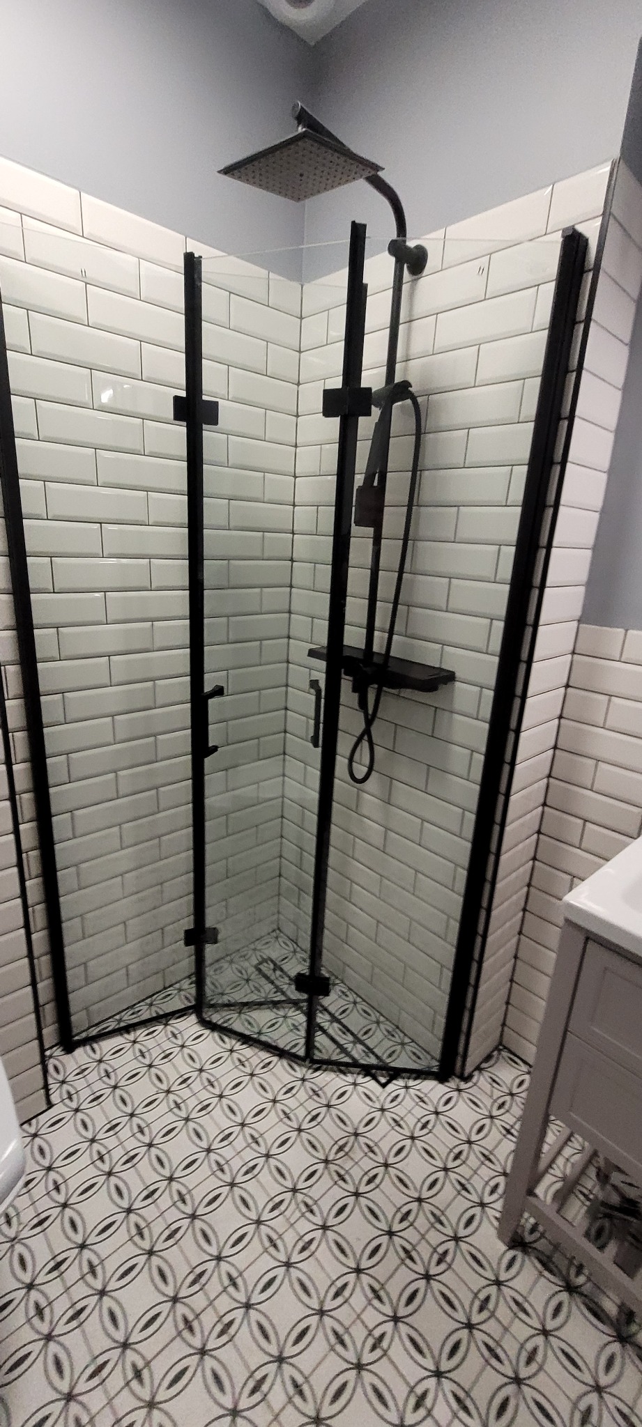 Nowoczesna kabina prysznicowa narożna z czarnymi profilami, białe płytki typu metro i wzorzysta podłoga, widok z góry.