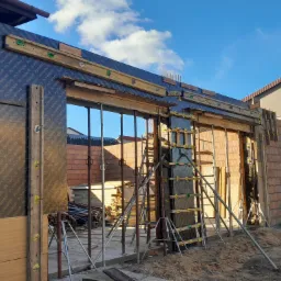 Szlifowanie betonu z użyciem szalunków systemowych i podpór teleskopowych, widok na nieukończone ściany z cegły i błękitne niebo.