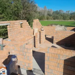 Wznoszone ściany parteru domu z czerwonej ceramiki, widoczne nadproże betonowe, paleta z bloczkami ceramicznymi i częściowo zapakowane bloczki, zielone drzewa w tle.