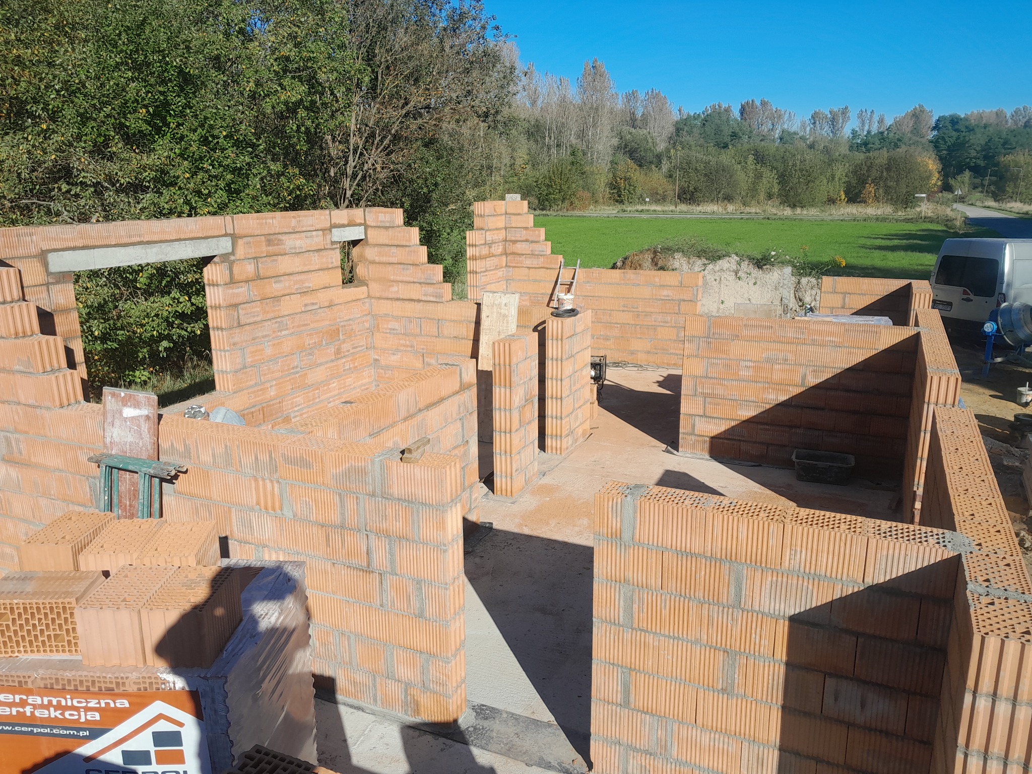 Wznoszone ściany parteru domu z czerwonej ceramiki, widoczne nadproże betonowe, paleta z bloczkami ceramicznymi i częściowo zapakowane bloczki, zielone drzewa w tle.