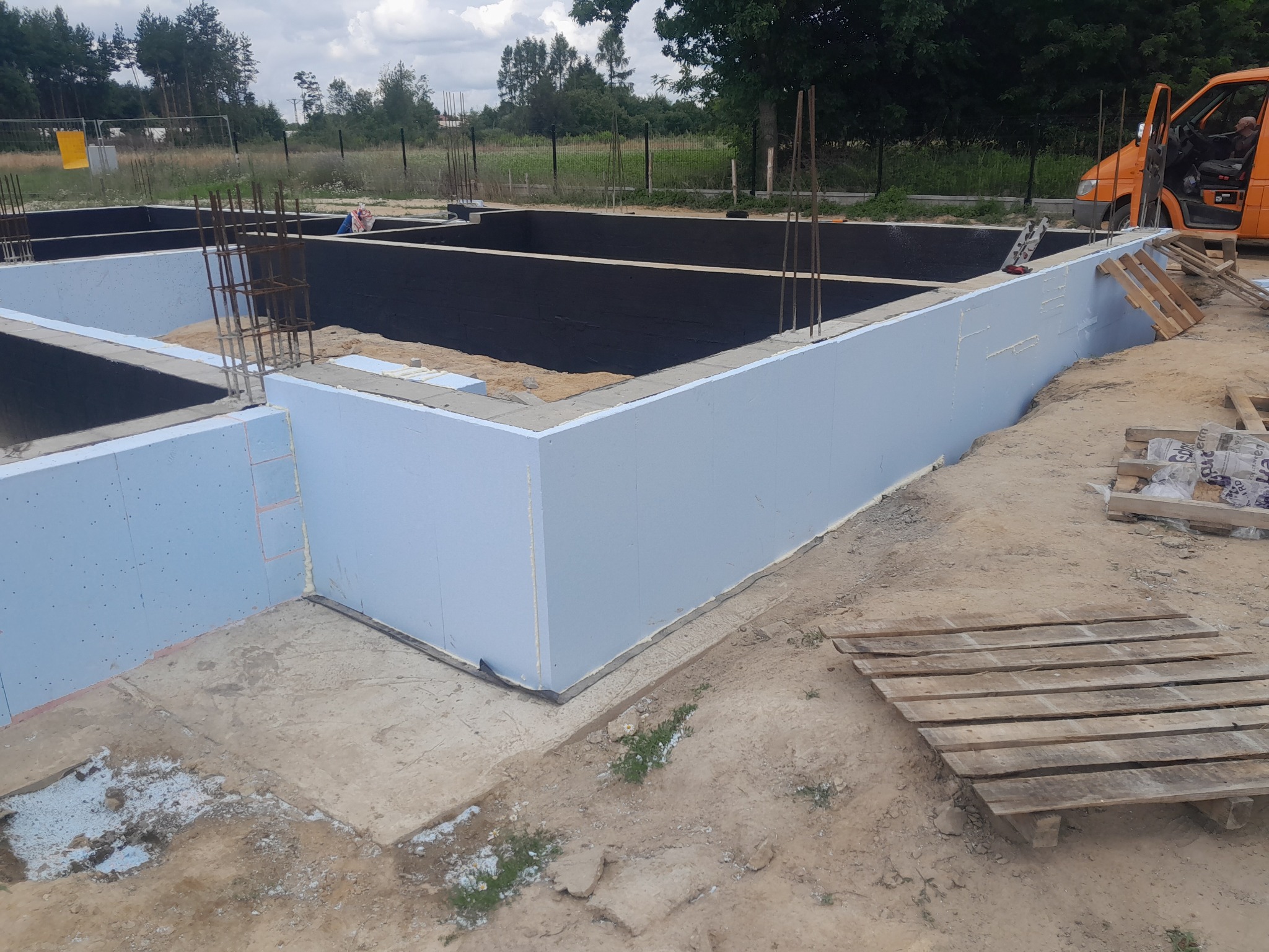 Ocieplone fundamenty budynku mieszkalnego z wystającymi prętami zbrojeniowymi, zabezpieczone czarną folią izolacyjną, widoczne palety i samochód dostawczy w tle.