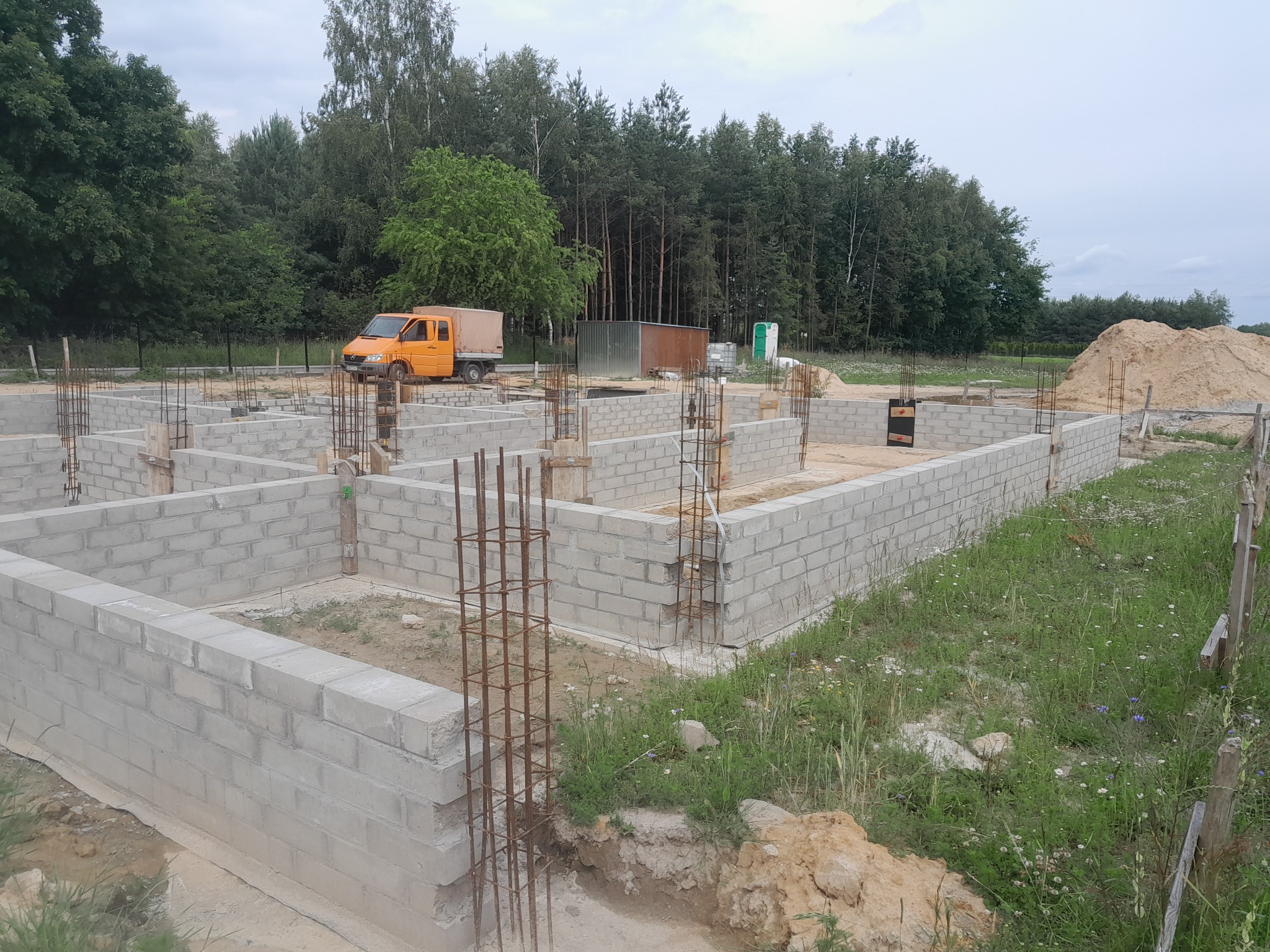 Fundamenty domu z bloczków betonowych, z wystającymi prętami zbrojeniowymi, pomarańczowy samochód dostawczy w tle.