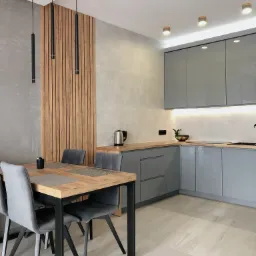 Nowoczesna kuchnia z szarymi frontami i drewnianym blatem, stół z metalowymi nogami i szarymi krzesłami, drewniana ścianka działowa z oświetleniem, minimalistyczne lampy wiszące.