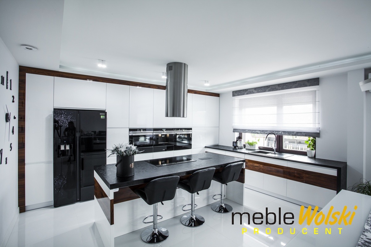 Nowoczesna, biała kuchnia z czarnym blatem i wyspą, srebrnym okapem cylindrycznym, zegarem ściennym i czarną lodówką z dekoracyjnym wzorem.