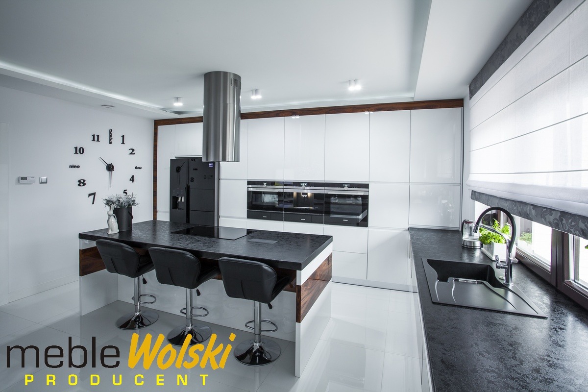 Nowoczesna, biała kuchnia z czarnym blatem i wyspą, chromowanymi hokerami, dużym zegarem ściennym i oknem z roletą rzymską.