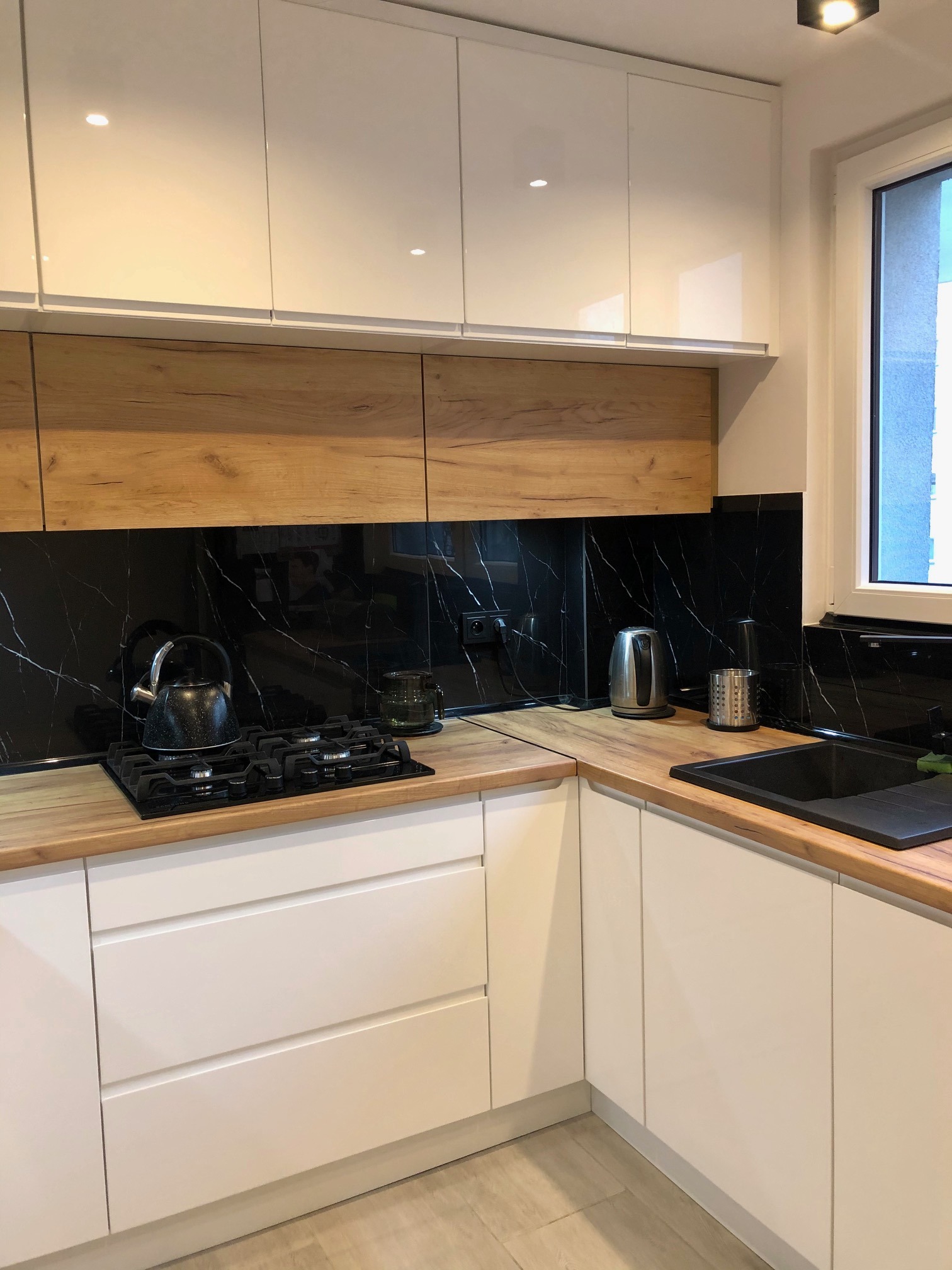 Nowoczesna kuchnia z białymi, lakierowanymi szafkami, drewnianymi blatami i czarnym panelem ściennym imitującym marmur, na blacie czajnik i płyta gazowa.