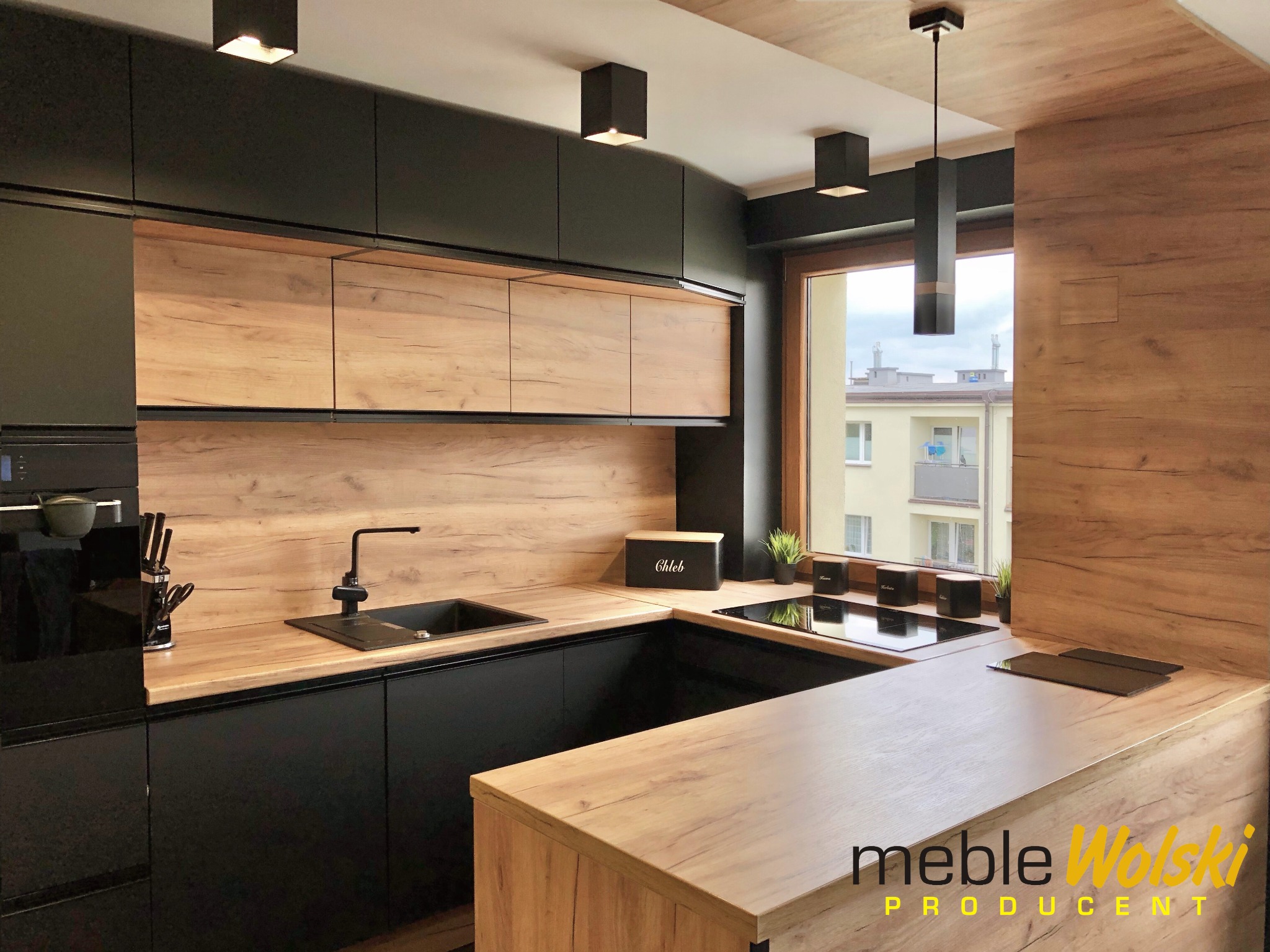 Nowoczesna kuchnia z czarnymi szafkami i drewnianymi blatami, oświetlenie punktowe i wiszące, widok na budynek z okna, z logo 'meble Wolski PRODUCENT' w prawym dolnym rogu.